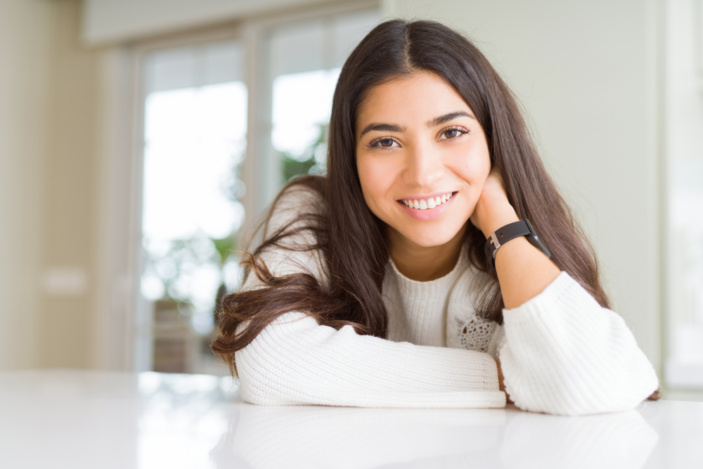 Woman with radiant smile relaxing at home – Endodontic Surgery A young woman with long hair smiles warmly while sitting at a bright table, reflecting renewed confidence and oral health – Endodontic Surgery.