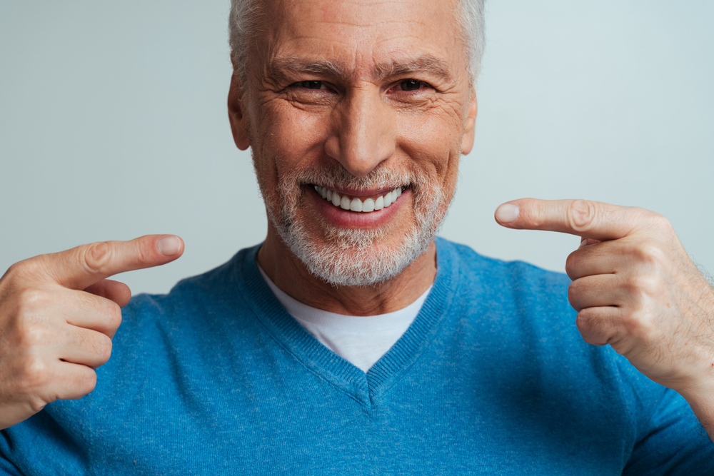 Mature man proudly showing his healthy smile – Root Canal Treatment A smiling older man in a blue sweater points to his bright, healthy teeth, expressing confidence and satisfaction – Root Canal Treatment.