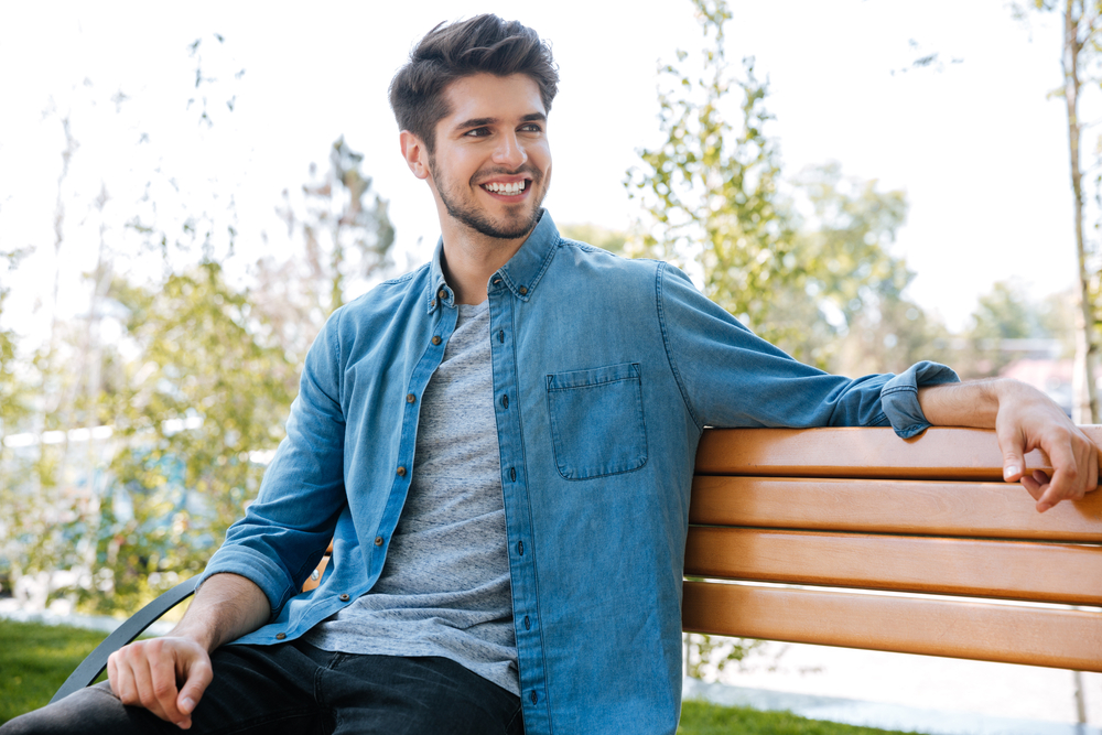 Smiling man sitting outdoors confidently – Traumatic Injuries A cheerful young man sits on a park bench smiling, symbolizing restored confidence and dental health after care – Traumatic Injuries.