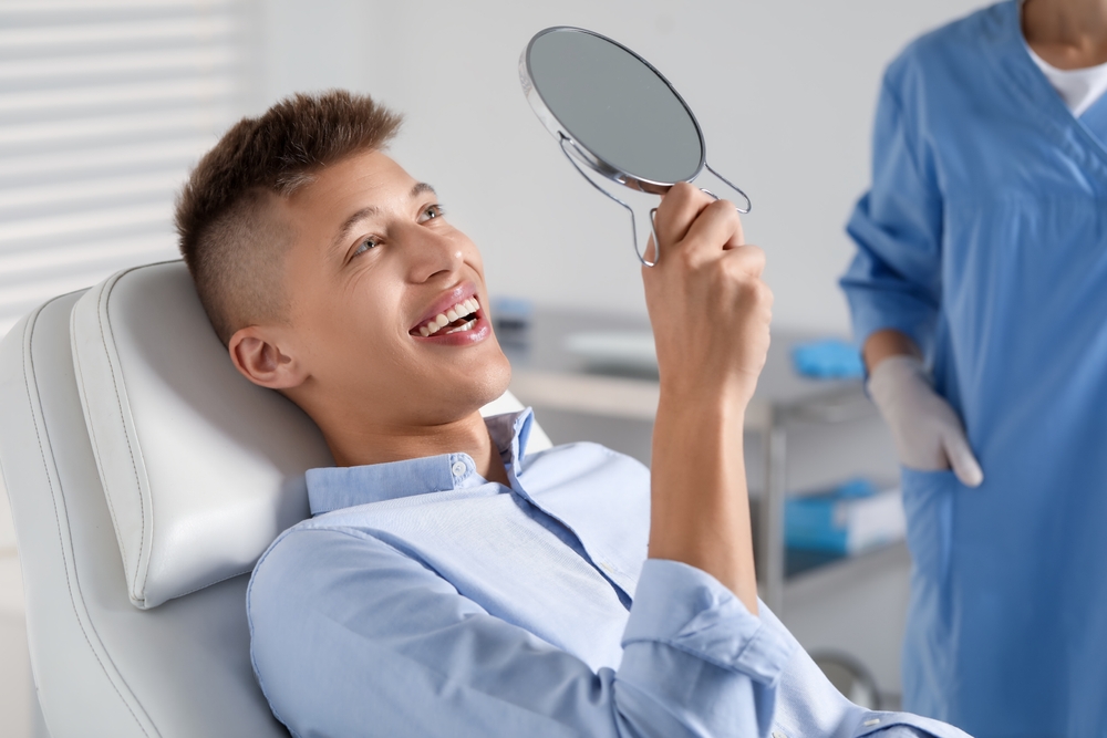 Young man smiling in dental chair holding mirror – Root Canal Bixby Knolls A young male patient smiles as he checks his reflection with a mirror after dental care, feeling satisfied and confident – Root Canal Bixby Knolls.