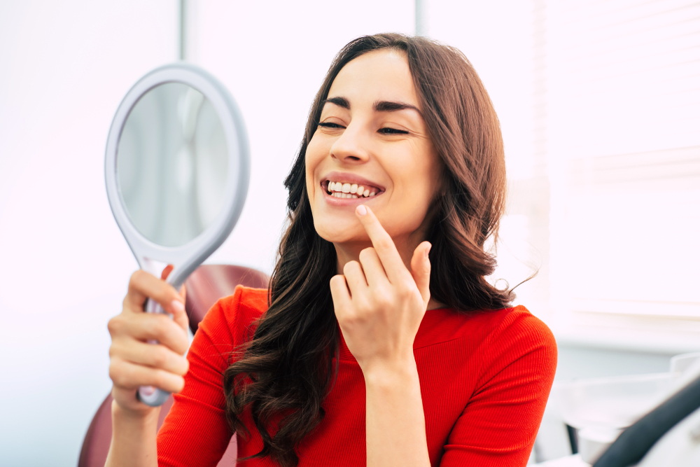 A cheerful woman in a red top examines her bright smile with a handheld mirror, showcasing satisfaction after treatment – Root Canal Retreatment.