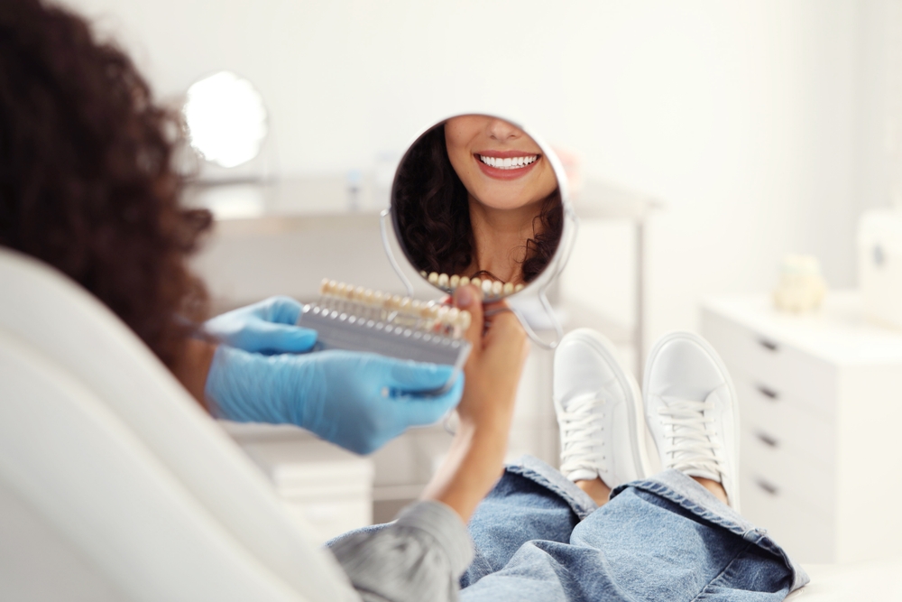 Patient comparing tooth shades in dental mirror – Non-Surgical Resorption Repair A woman sits in a dental chair holding a mirror while matching tooth shades, showcasing restored dental aesthetics – Non-Surgical Resorption Repair.