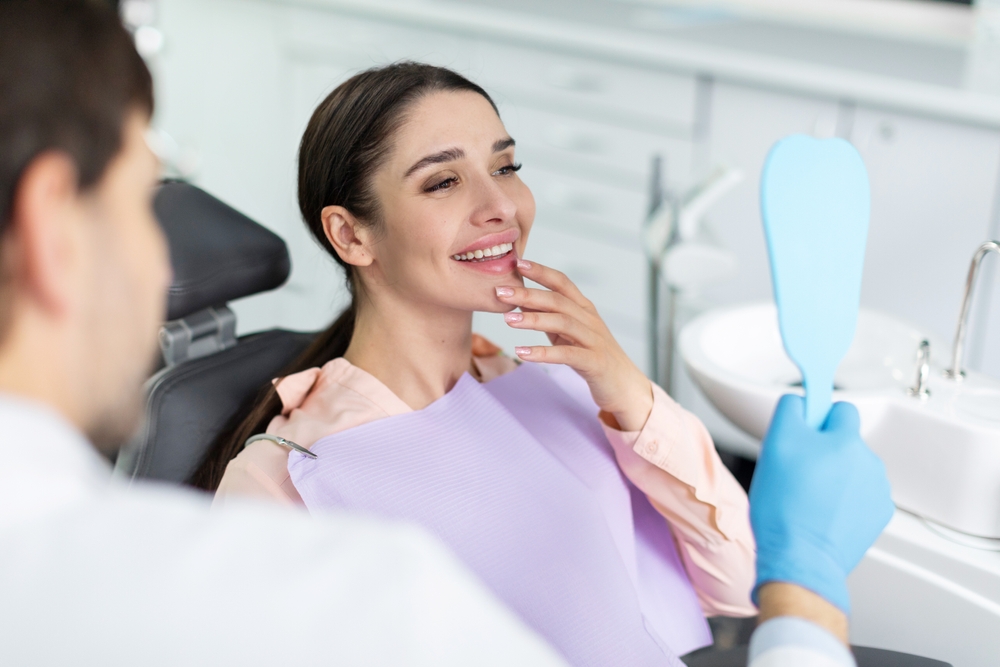 Woman checking her reflection after dental visit – Gentlewave A smiling woman looks into a handheld mirror at the dental clinic, admiring her restored and healthy smile – Gentlewave.