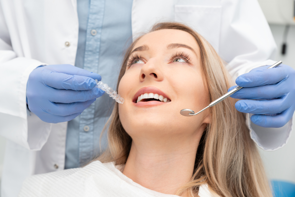 Dentist showing clear aligner to patient – Root Canal Treatment A female patient looks up at her dentist holding a clear aligner and dental tools, symbolizing precision and advanced care – Root Canal Treatment.
