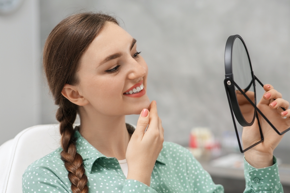 Woman admiring her smile in the mirror after dental care – Cracked Teeth Diagnosis and Treatment A young woman with a braid smiles while examining her reflection with a handheld mirror, highlighting confidence and satisfaction following cracked teeth diagnosis and treatment.