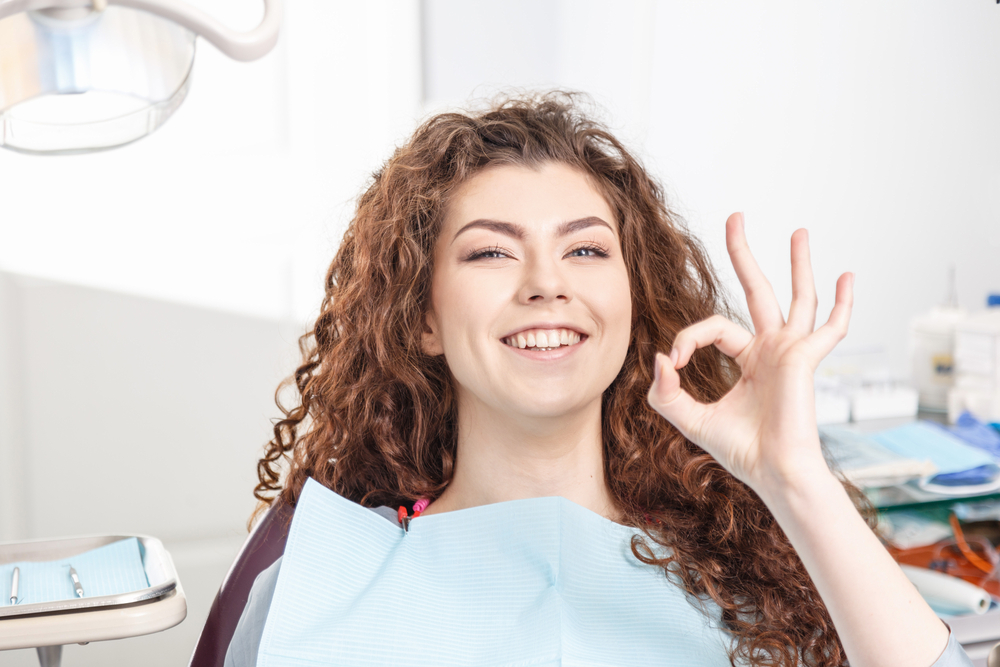 Happy patient giving OK sign after dental visit – Cracked Teeth Diagnosis and Treatment A smiling woman with curly hair gives an OK hand gesture while sitting in the dental chair, expressing satisfaction and relief after receiving expert cracked teeth diagnosis and treatment.