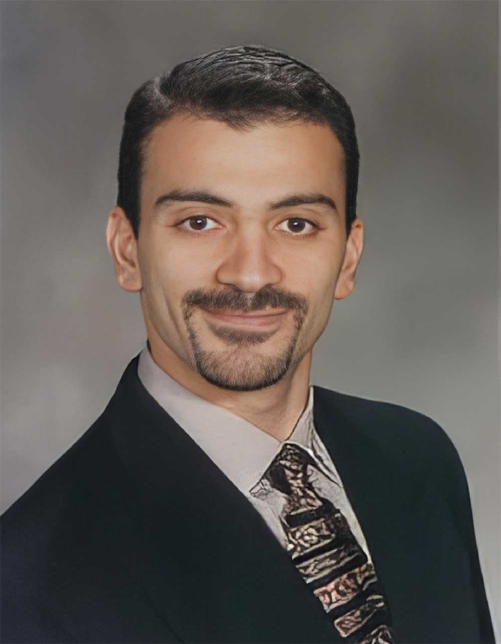 Dr. Ahmed Sadek - Marina Endodontics A professional headshot of Dr. Ahmed Sadek, wearing a dark suit, light dress shirt, and patterned tie, posed against a neutral gray background. - Marina Endodontics