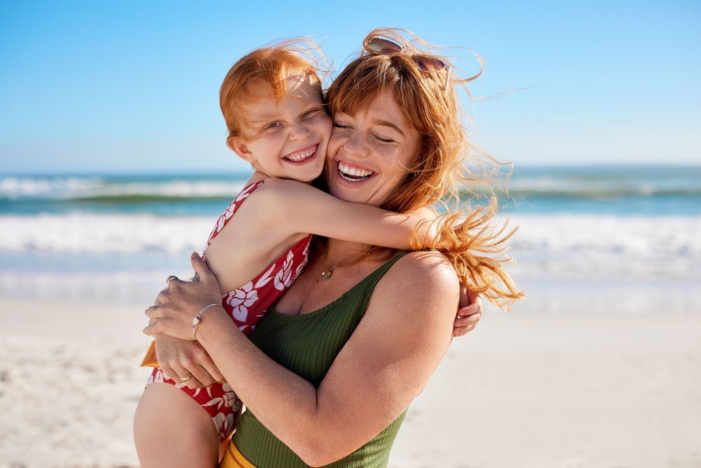Smiling woman hugs a young girl on a sunny beach. - Marina Endodontics A happy woman and child embrace on the sand with the ocean in the background. - Marina Endodontics