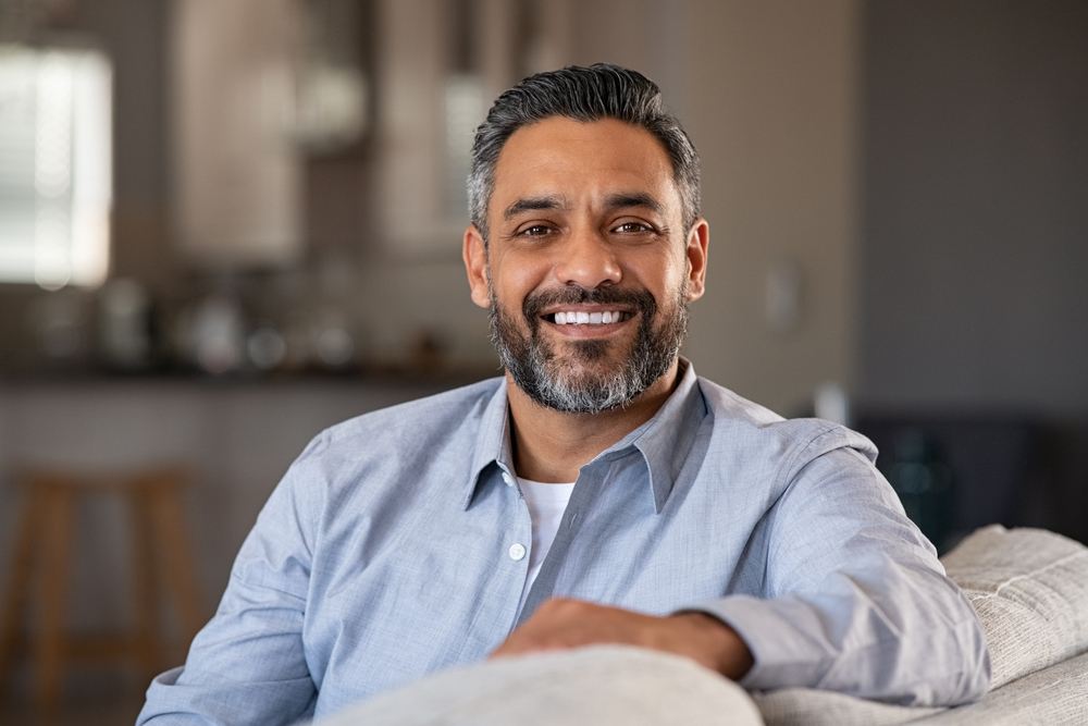 Confident man smiling at home – Gentlewave A smiling man with salt-and-pepper hair sits comfortably on a couch, showing confidence and comfort after advanced dental treatment – Gentlewave.