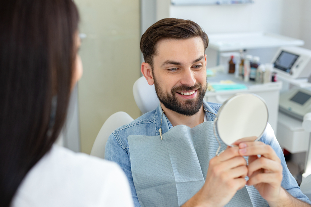 A male patient sits in a dental chair smiling while looking at his reflection in a mirror, appreciating his restored smile – Root Canal Retreatment.