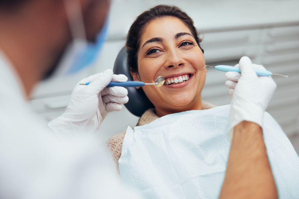 Root Canal Treatment - Marina Endodontics A smiling patient sits in a dental chair while a dentist prepares to perform a root canal procedure. The dentist, wearing gloves and a mask, holds dental instruments including a mirror and scaler, while the patient looks relaxed and comfortable. - Marina Endodontics