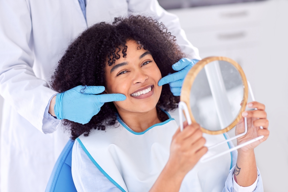 Patient admiring her bright smile in dental mirror – Gentlewave A happy woman sits in a dental chair holding a mirror, while her dentist points out her beautiful results after treatment – Gentlewave.