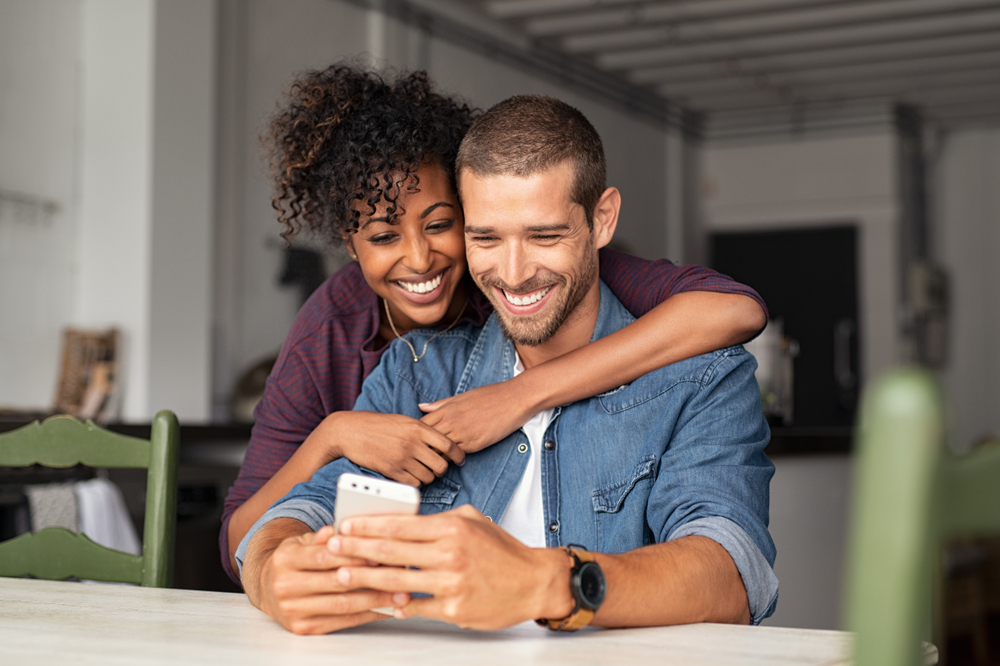 Happy couple smiling while looking at phone – Endodontic Surgery A cheerful couple smiles together while viewing something on a smartphone, representing restored confidence and comfort after treatment – Endodontic Surgery.